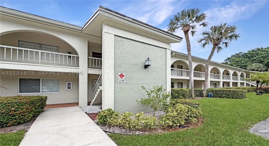 Multi-story stucco exterior featuring arched balcony railings, concrete walkways, built-in wall-mounted lighting, and landscaped shrubbery