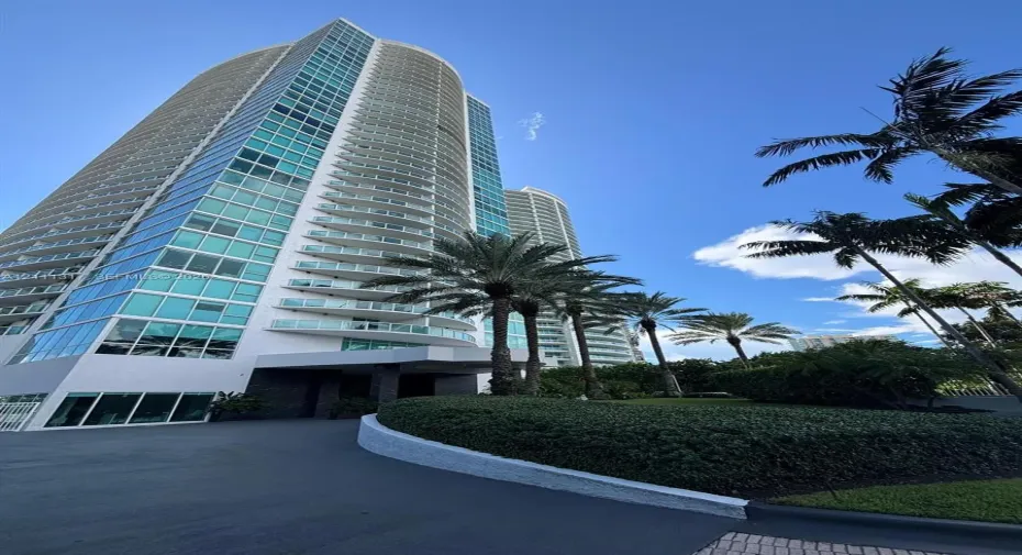 High-rise architecture featuring a glass facade, curved structural balconies, a porte-cochère entry, and mature landscaping