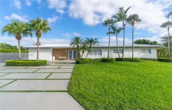 Single-story residence featuring a standing-seam metal roof, horizontal wood-panel siding, a concrete paver driveway with integrated turf, and professional landscaping with mature palm trees