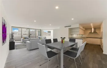 Open-concept living space featuring wood-finish flooring, recessed lighting, floor-to-ceiling glass sliding doors, and a kitchen equipped with flat-panel cabinetry, a stainless steel range hood, and solid-surface countertops