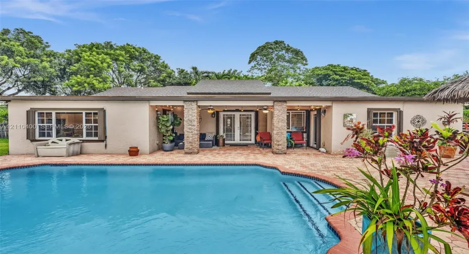 Stucco exterior featuring a covered patio with stone-finished pillars, a paver deck, and an in-ground swimming pool with steps