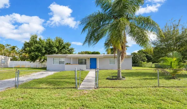 Single-story exterior featuring a concrete walkway, chain-link perimeter fencing, and mature palm landscaping