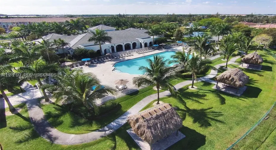 Community recreation area featuring a large swimming pool, concrete sun deck, and multiple thatched-roof gazebos set within a landscaped lawn