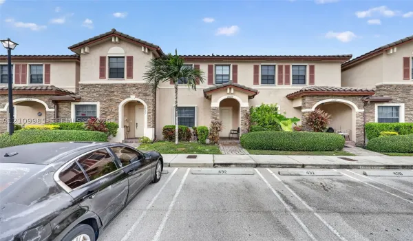 Two-story Mediterranean-style townhome exterior featuring stucco and stone veneer cladding, arched entryway accents, tile roofing, and assigned parking spaces