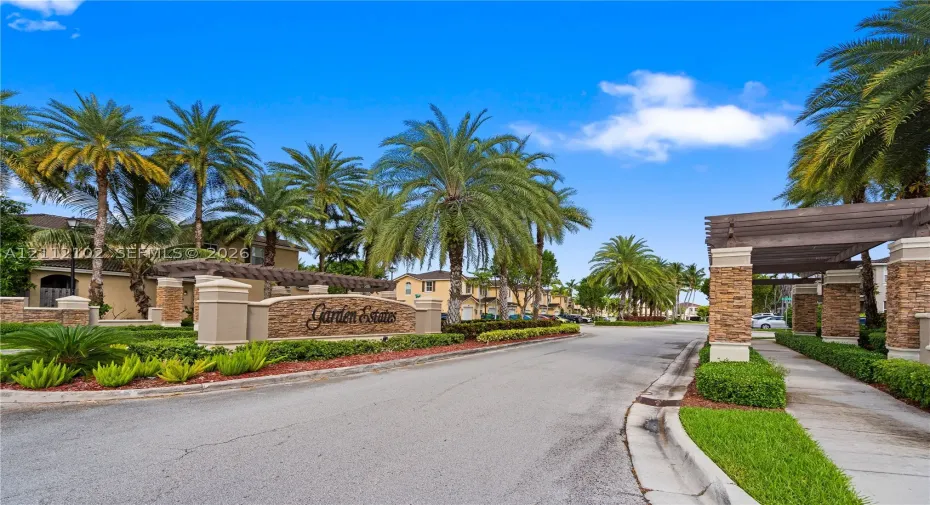 Community entrance featuring stone-clad pillars, a wood-beam pergola, mature palm trees, and manicured landscaping