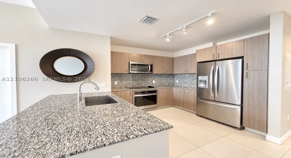 Kitchen featuring flat-panel wood-finish cabinetry, speckled stone countertops, stainless steel appliances, a center island with an undermount sink, and tile flooring