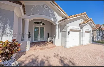 Stucco exterior featuring an arched entryway with dual columns, a clay tile roof, herringbone-pattern brick paver driveway, and a double-door glass entrance