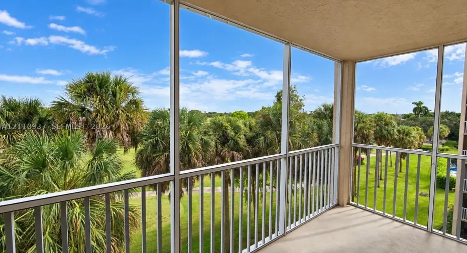 Screened balcony featuring metal railings, overhead soffit, and concrete flooring, overlooking a landscape of mature palms and expansive lawn