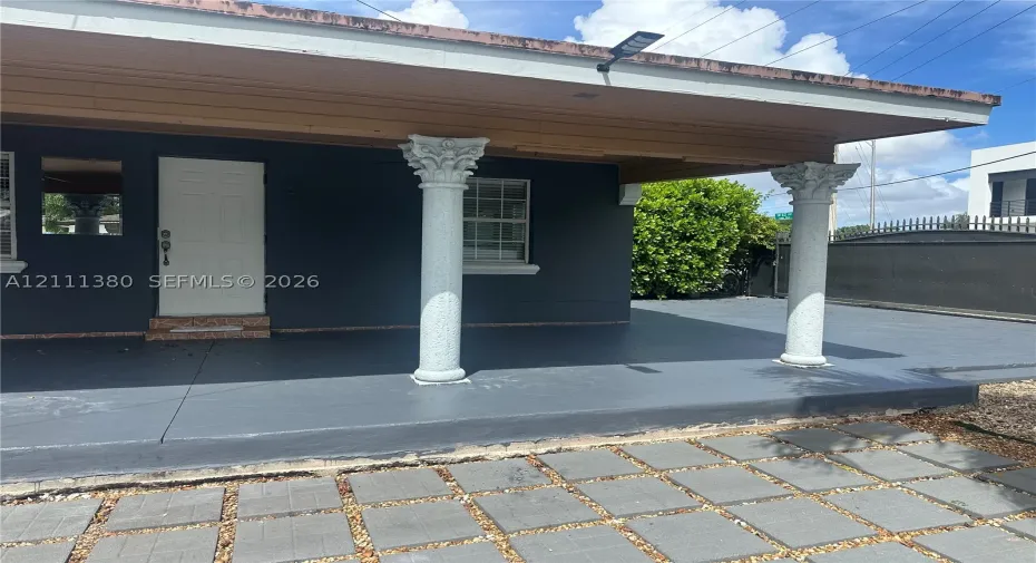 Covered front porch featuring decorative columns with ornate capitals, a paved hardscape entry, and a solid exterior door