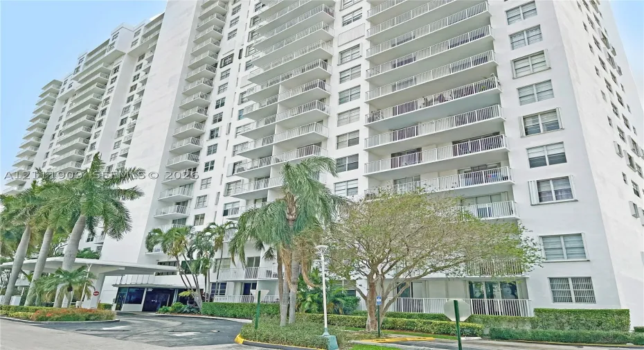 High-rise multi-story building featuring tiered private balconies, an expansive concrete motor court, a covered entryway portico, and mature tropical landscaping