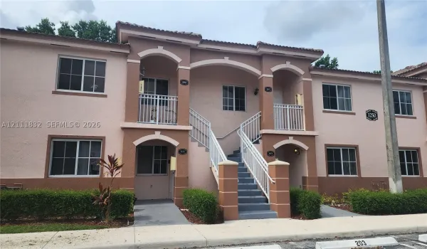 Stucco exterior featuring arched entryways, concrete stairs with white metal railings, tiled roofline, and manicured shrubbery