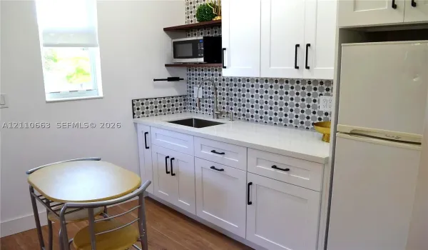 White Shaker-style cabinetry featuring matte black hardware, stone-finish countertops, patterned tile backsplash, stainless steel sink with gooseneck faucet, and wood-finish flooring