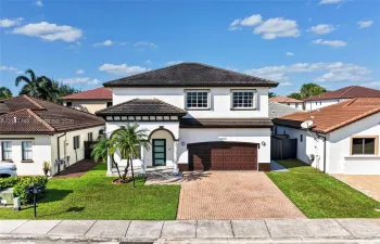 Two-story stucco exterior featuring a multi-hipped tile roof, paver driveway, covered entryway with arched architectural detailing, and a multi-panel garage door
