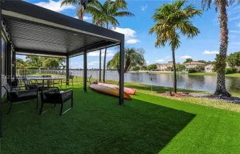 Covered patio featuring a dark metal pergola, synthetic grass lawn, perimeter fencing, and water frontage with mature palm trees