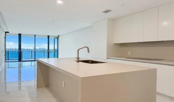 Modern kitchen featuring a central island with a waterfall countertop, integrated stainless steel sink, polished stone-finish flooring, floor-to-ceiling glass sliding doors, and minimalist cabinetry