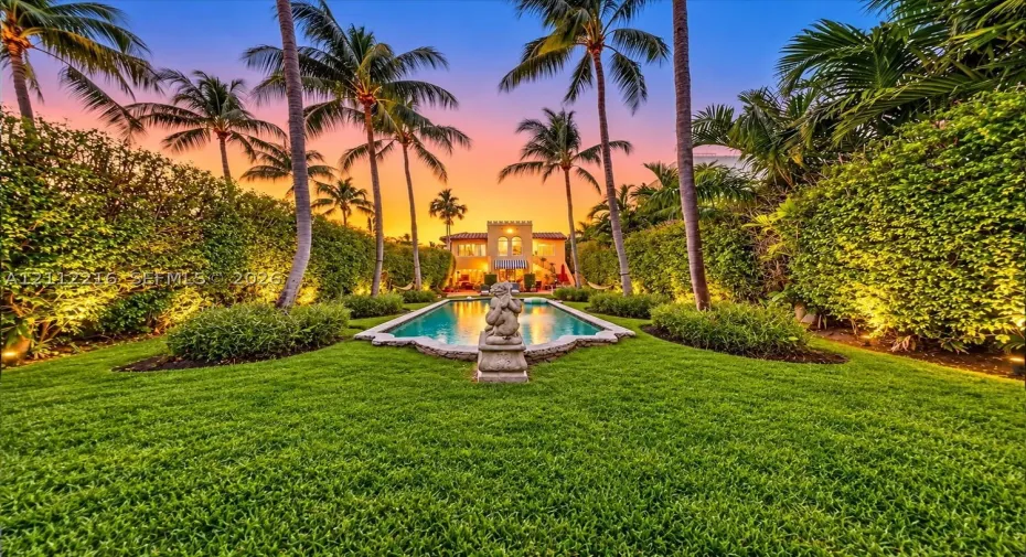 Expansive backyard featuring a rectangular swimming pool, stone sculpture, manicured lawn, and mature palm trees framed by high-density hedges