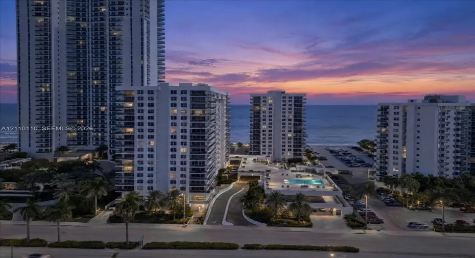 High-rise coastal residential structures featuring private balconies, an elevated swimming pool, and mature palm landscaping with a direct oceanfront backdrop