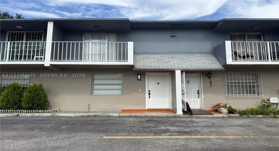 Two-story building exterior featuring upper-level balconies with white railings, louvered windows with security bars, and dual ground-level entry doors