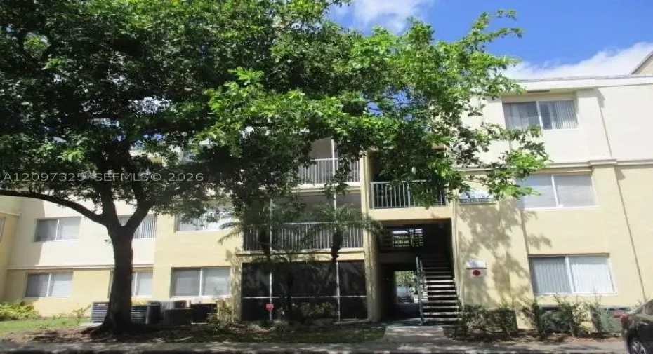 Multi-story exterior featuring a light-toned stucco facade, screened balconies with metal railings, and a concrete staircase