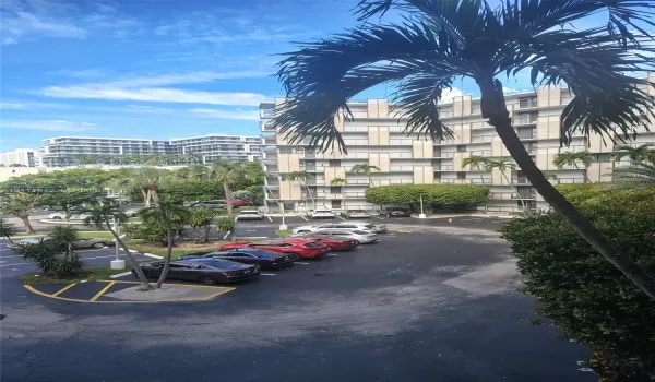Multi-story building featuring concrete balconies, landscaped grounds with palm trees, and paved surface parking