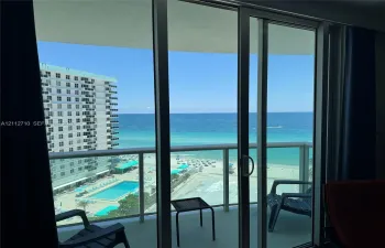 Floor-to-ceiling glass sliding doors opening to a private balcony with glass panel railings, featuring expansive ocean scenery