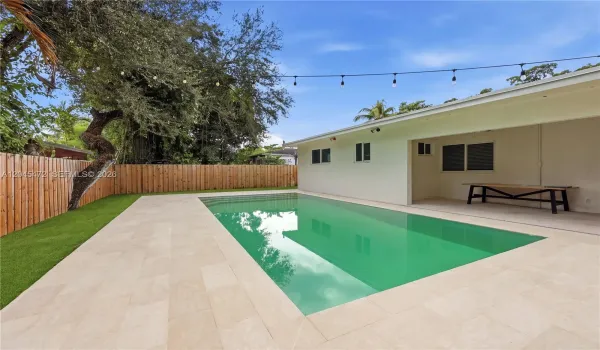 In-ground pool with stone paver decking, enclosed by a wood privacy fence and featuring a covered patio area with synthetic lawn