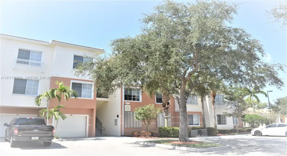 Multi-story residential building featuring a stucco exterior, integrated garage, private balconies with railings, and mature landscaping