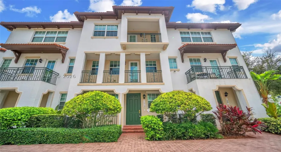 Stucco multi-story exterior featuring a tile roof, multiple wrought-iron balconies, arched entryway detailing, and brick-paver hardscaping