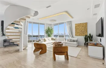 Open-concept living space featuring a sculptural spiral staircase with wood-tread steps, polished stone flooring, recessed ceiling lighting, and floor-to-ceiling glass panel walls