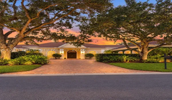 Single-story exterior featuring arched windows, a covered entryway with structural columns, and a paver driveway, complemented by mature canopy trees and manicured landscaping