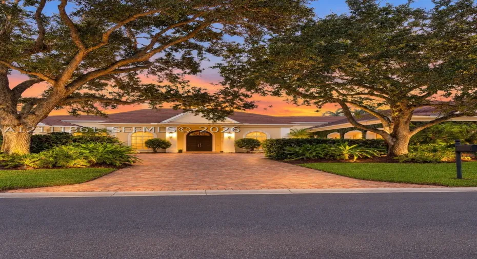 Single-story exterior featuring arched windows, a covered entryway with structural columns, and a paver driveway, complemented by mature canopy trees and manicured landscaping
