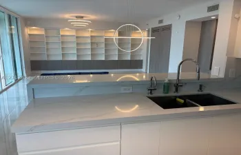 Kitchen island featuring white countertops, a double-basin sink with a gooseneck faucet, built-in shelving with integrated lighting, and a modern circular pendant light fixture