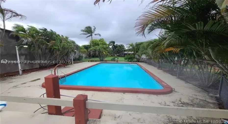 Rectangular in-ground swimming pool featuring a terra cotta border, metal handrail, concrete pool deck, perimeter fencing, and mature palm trees