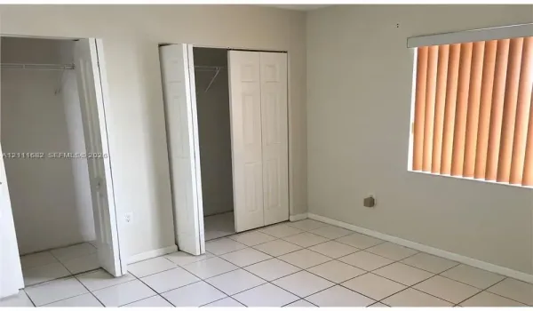 Interior space featuring light-colored tile flooring, two closets with white bi-fold doors, and a window fitted with vertical blinds