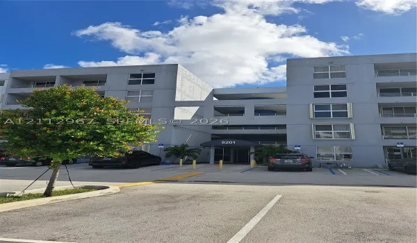 Multi-story residential exterior featuring concrete facade, private balconies, covered building entrance with awning, and surface parking