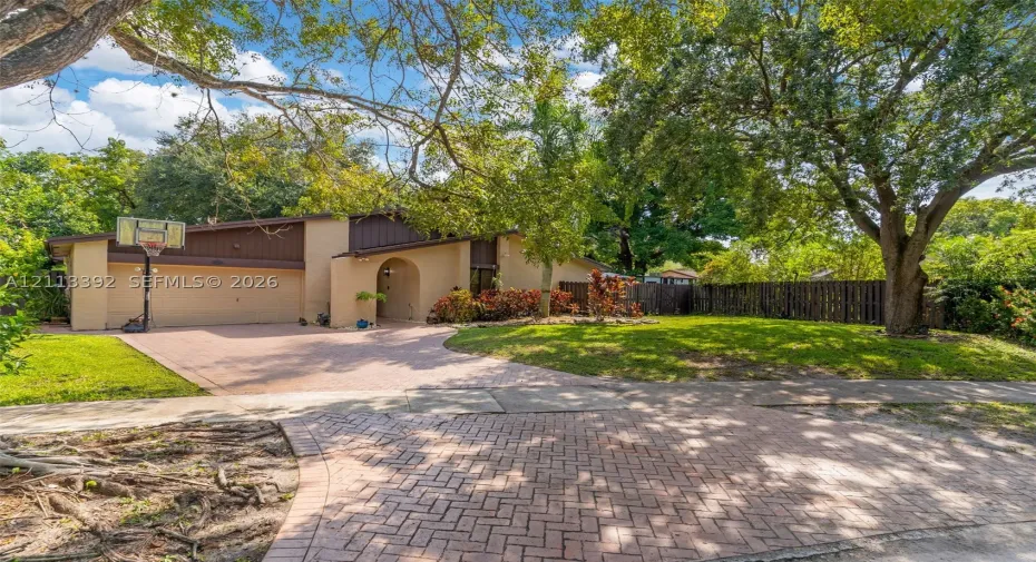 Exterior elevation featuring a multi-tonal facade, paved brick driveway, attached two-car garage, mature landscaping, and fenced perimeter