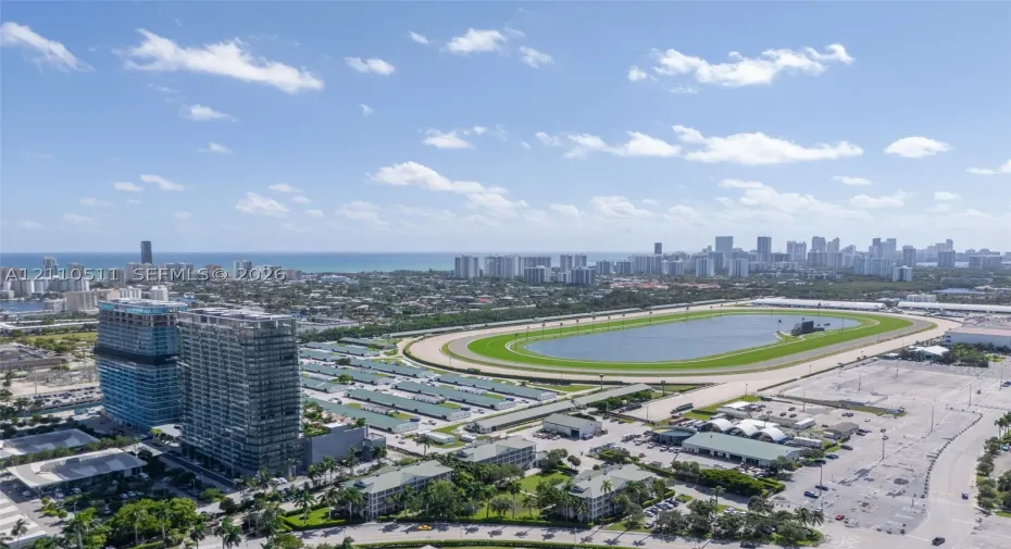 Aerial perspective featuring a multi-story glass-facade residential building, a large-scale race track, and a coastal skyline backdrop