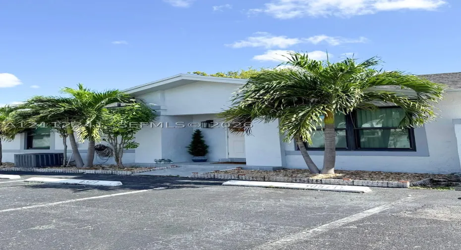 Stucco exterior featuring dual-pane windows with dark trim, a covered entryway, paved walkway, and landscaping including mature palm trees