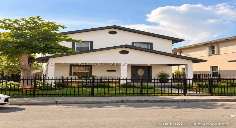 Two-story exterior featuring light-toned stucco facade, covered front porch with square columns, dark-trimmed windows, and decorative metal perimeter fencing