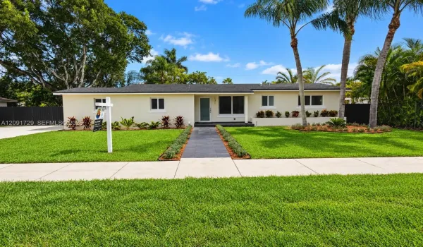 Single-story residence with a light-colored stucco exterior, dark shingle roof, centered entryway, stone-tiled walkway, and mature palm trees