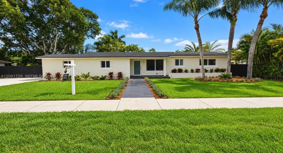 Single-story residence with a light-colored stucco exterior, dark shingle roof, centered entryway, stone-tiled walkway, and mature palm trees