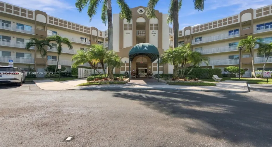 Multi-story stucco exterior featuring a covered entryway with a green metal awning, private balconies, and landscaped grounds with mature palm trees