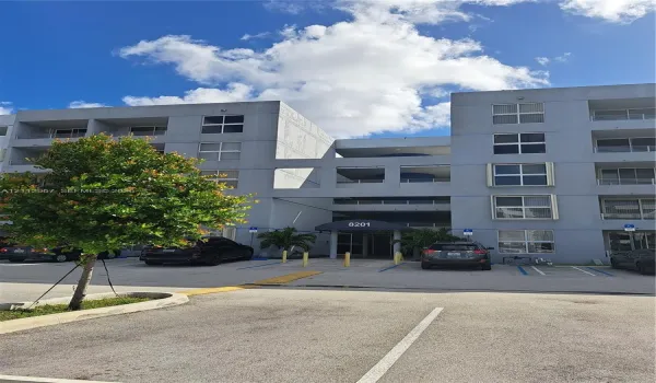 Multi-story residential exterior featuring concrete facade, private balconies, covered building entrance with awning, and surface parking