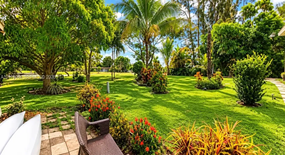 Expansive backyard featuring mature shade trees, established ornamental landscaping, palm trees, and a paved stone patio