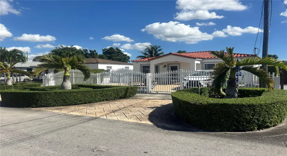 Exterior featuring a tile roof, paved driveway, and decorative white metal fencing surrounding the perimeter, complemented by manicured shrubbery