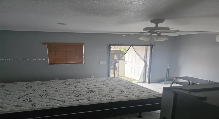 Neutral-toned interior space featuring light-colored tile flooring, a multi-blade ceiling fan with integrated lighting, a single-hung window with wood-finish blinds, and a paneled exterior door