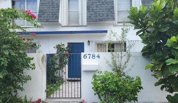 Multi-story exterior featuring a shingled mansard roof, a gated courtyard entry, a solid panel front door, and white stucco siding