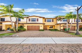 Two-story townhouse with warm hues, brown accents, single-car garage, and brick driveway. Lush greenery and palm trees add tropical charm. Inviting suburban home.