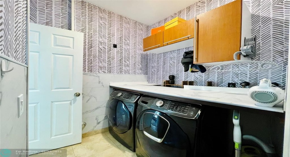 Modern laundry room with two black Samsung front-loading washing machines, a white marble-like countertop, and wooden cabinets mounted above. The walls feature black and white herringbone patterned wallpaper, and a ceiling light illuminates the space. The floor is tiled, and there is a white door to the right. Various utility items are neatly organized, and the room has a clean, stylish appearance.